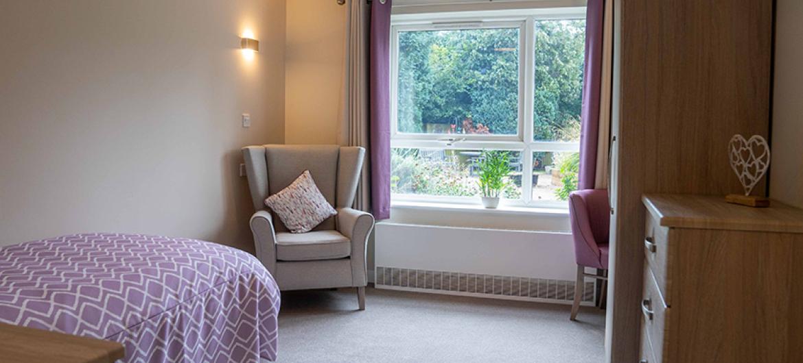 Spacious single bed room with large window facing the garden area. A chesterfield arm chair sits to the window, the room is finished with a wooden wardrobe and matching chest of draw set