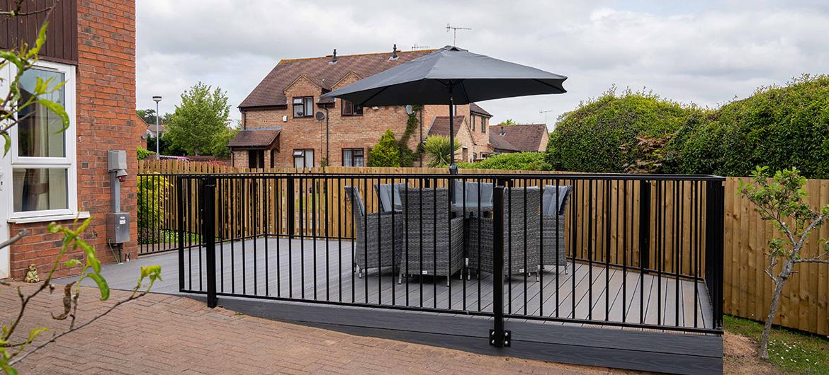 Raised patio area surrounded by iron safety railings and a large six person wicker outdoor dining table and chairs with covered parasol